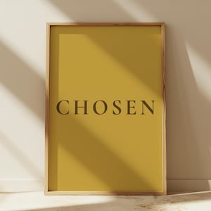 May include: A rectangular wall art print in a light wood frame featuring a solid mustard yellow background with the word CHOSEN printed in a dark brown serif font centered horizontally in the middle of the composition.