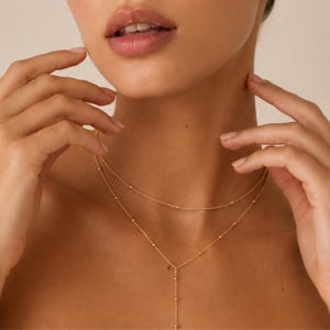 May include: A delicate gold-toned layered necklace featuring two thin chains adorned with small, evenly spaced metallic beads. The upper chain sits closer to the neck, while the lower chain features a vertical drop element in a Y-shape design.
