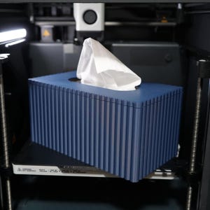 Peut inclure: Un boîtier à mouchoirs rectangulaire bleu foncé avec une texture nervurée verticale. Le boîtier présente une ouverture ovale centrale d'où dépasse un mouchoir blanc. Il est placé dans une imprimante 3D avec un volume de 256x256mm.