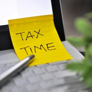 Peut inclure: Un post-it jaune collé sur un clavier d'ordinateur portable affiche les mots "TAX TIME" en noir. Un stylo argenté repose sur le clavier, et une plante verte floue est en arrière-plan. L'image suggère un thème fiscal ou financier.