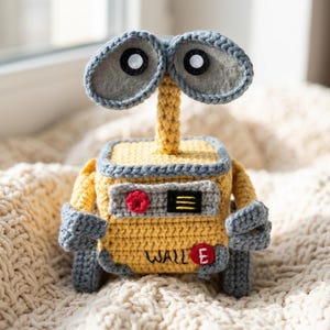 May include: A crochet toy of the robot WALL-E, featuring a yellow box-shaped body, grey arms, and two large oval eyes on a central stalk. The front panel displays a red button, a black rectangular detail with yellow lines, and the text WALL-E.