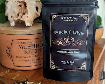 May include: A black pouch labelled Wild & Whimsy Witches' Elixir Samhain Seasonal Blend Herbal Tea Blend Loose Leaf 25g. A wooden spoon holds a mixture of dried blue flowers, green herbs, and light-coloured pieces of dried fruit.