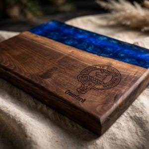 May include: A rectangular wooden serving board featuring a dark brown wood grain section and a vibrant blue resin inlay. The wood surface is engraved with a circular crest depicting a stag and the text "TUTUM TE ROBORE REDDAM" and "Crawford".