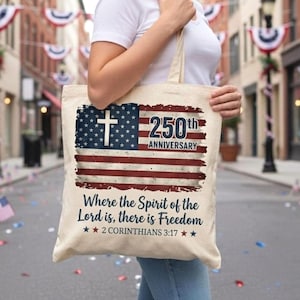 Puede incluir: Un bolso tote beige con un diseño de bandera estadounidense desgastada, una cruz blanca y el texto "250th Anniversary". El bolso también presenta el texto "Where the Spirit of the Lord is, there is Freedom" y "2 Corinthians 3:17."