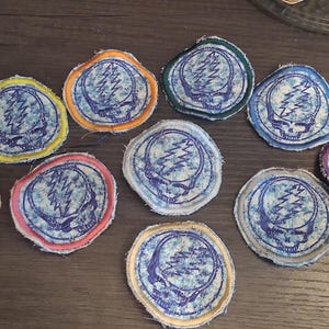 Könnte beinhalten: Eine Sammlung von zehn bunten, runden Aufnähern. Jeder Aufnäher zeigt ein blau-weißes Design eines Blitzes über einem Totenkopf. Die Aufnäher haben verschiedenfarbige Ränder, darunter rot, gelb, orange und lila.