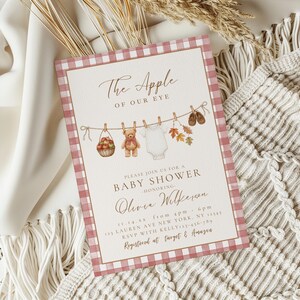 May include: A rectangular baby shower invitation card with a pink and white gingham border. The text reads The Apple of Our Eye, Please Join Us for a Baby Shower Honoring Olivia Wilkerson, 11.14.xx, 123 Lauren Ave New York, NY 12345, RSVP with Kelly 123-456-789, Registered at Target & Amazon. Illustrations include a basket of apples, a teddy bear, a white onesie, autumn leaves, and baby shoes hanging on a clothesline.