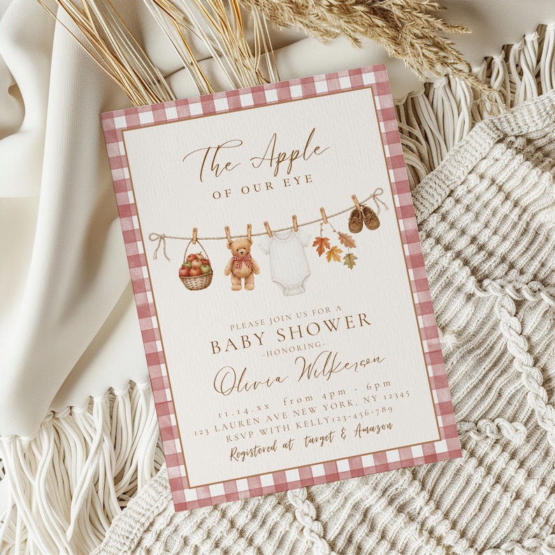 May include: A rectangular baby shower invitation card with a pink and white gingham border. The text reads The Apple of Our Eye, Please Join Us for a Baby Shower Honoring Olivia Wilkerson, 11.14.xx, 123 Lauren Ave New York, NY 12345, RSVP with Kelly 123-456-789, Registered at Target & Amazon. Illustrations include a basket of apples, a teddy bear, a white onesie, autumn leaves, and baby shoes hanging on a clothesline.