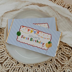 Puede incluir: Dos barras de chocolate rectangulares envueltas en papel a rayas azules y blancas. Las etiquetas presentan un rectángulo blanco festoneado con banderines coloridos, globos y el texto "Oliver's First Birthday" en una fuente multicolor.
