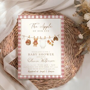 May include: A rectangular baby shower invitation card with a red and white gingham border. The text reads The Apple OF OUR EYE, BABY SHOWER, honoring Olivia Wilkerson, with event details, and illustrations of a basket of apples, a teddy bear, a onesie, autumn leaves, and baby shoes hanging on a clothesline.