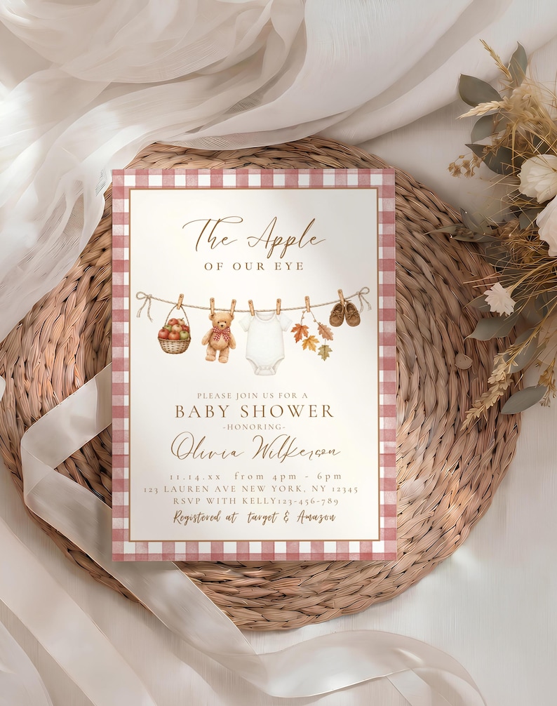May include: A rectangular baby shower invitation card with a red and white gingham border. The text reads The Apple OF OUR EYE, BABY SHOWER, honoring Olivia Wilkerson, with event details, and illustrations of a basket of apples, a teddy bear, a onesie, autumn leaves, and baby shoes hanging on a clothesline.