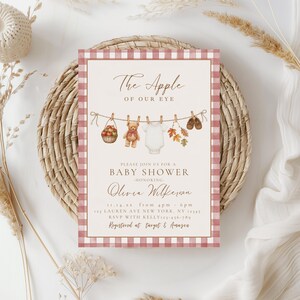 May include: A rectangular baby shower invitation card with a pink and white gingham border. The center features a clothesline with a basket of apples, a teddy bear, a white onesie, autumn leaves, and baby shoes. Text reads: The Apple of Our Eye, Baby Shower, Olivia Wilkerson, 11.14.xx, 4pm-6pm, 123 Lauren Ave New York, NY 12345, RSVP with Kelly 123-456-789, Registered at Target & Amazon.