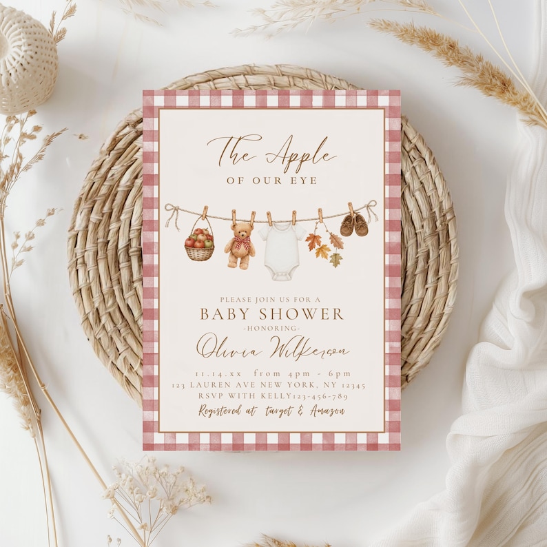 May include: A rectangular baby shower invitation card with a pink and white gingham border. The center features a clothesline with a basket of apples, a teddy bear, a white onesie, autumn leaves, and baby shoes. Text reads: The Apple of Our Eye, Baby Shower, Olivia Wilkerson, 11.14.xx, 4pm-6pm, 123 Lauren Ave New York, NY 12345, RSVP with Kelly 123-456-789, Registered at Target & Amazon.