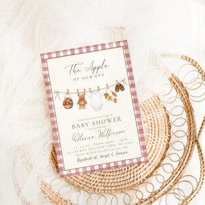 May include: A rectangular baby shower invitation card featuring a red and white gingham border. The center displays a clothesline with a basket of apples, a teddy bear, a white onesie, autumn leaves, and baby shoes, with the text The Apple of Our Eye.