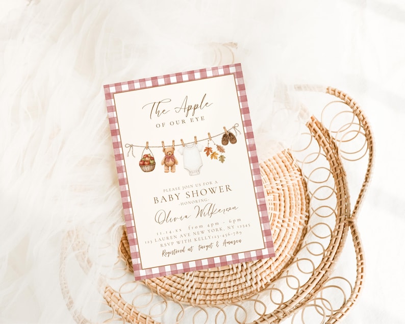 May include: A rectangular baby shower invitation card featuring a red and white gingham border. The center displays a clothesline with a basket of apples, a teddy bear, a white onesie, autumn leaves, and baby shoes, with the text The Apple of Our Eye.