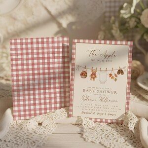 May include: A baby shower invitation card featuring a red and white gingham border and a central illustration of a clothesline with a basket of apples, a teddy bear, a white onesie, autumn leaves, and baby shoes, with printed event details.