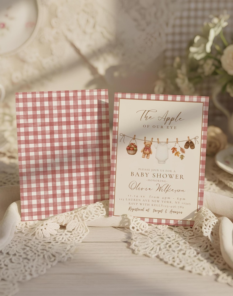May include: A baby shower invitation card featuring a red and white gingham border and a central illustration of a clothesline with a basket of apples, a teddy bear, a white onesie, autumn leaves, and baby shoes, with printed event details.