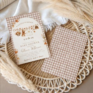 Może przedstawiać: Zaproszenie na baby shower z brązowo-białą kratką vichy, łukowatą ramką oraz ilustracjami misia, ubranek dziecięcych i jesiennych liści. Tekst zawiera napis A Little Pumpkin is on the way, Baby Shower oraz szczegóły dotyczące wydarzenia.