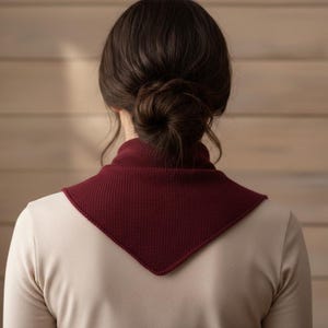 May include: A burgundy knit cowl neck warmer. The triangular scarf is worn over a cream-coloured long-sleeved shirt. The background is a light brown wooden wall.