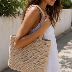 May include: A beige tote bag crafted from woven material with a rectangular shape and two sturdy shoulder straps. The bag features a consistent, tight crochet-like texture throughout its surface, providing a structured yet flexible appearance.
