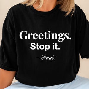 以下が含まれることがあります： 前面に白い文字がプリントされた黒の半袖Tシャツ。「Greetings. Stop it. — Paul.」というテキストが書かれています。Tシャツはクルーネックでゆったりとしたフィット感があり、グラフィックが胸の中央に配置されています。
