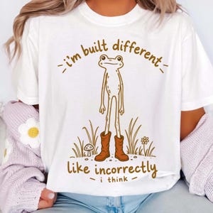 Puede incluir: Una camiseta blanca con una ilustración de líneas marrones de una rana alta y delgada con botas marrones. El texto superior dice i'm built different y el texto inferior dice like incorrectly i think, rodeado de hierba y una seta.