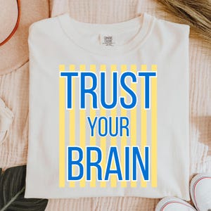 May include: Cream-colored sweatshirt with the words "TRUST YOUR BRAIN" in blue, set against a backdrop of vertical yellow stripes. The shirt is made of a soft material and has a crew neck.
