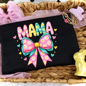以下が含まれることがあります： カラフルな文字で「MAMA」と書かれた黒いTシャツと大きなリボンのグラフィック。リボンはピンクとティールで、デイジーのアクセントが付いています。シャツはハートと黄色のアクセントで飾られています。
