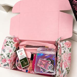 May include: A pink gift box filled with various items, including a pen, perfume, stickers, and a small box with a flamingo design. The box is lined with floral tissue paper.