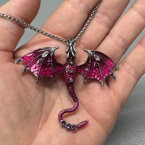 May include: A silver-toned dragon pendant necklace with a magenta and pink enamel finish. The dragon has outstretched wings with crystal accents and a curved tail. The necklace is displayed on a hand.