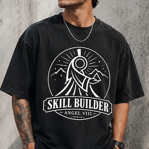 May include: A black short-sleeve t-shirt featuring a white circular graphic on the chest. The design depicts a stylized figure holding a pyramid with mountains in the background. The text reads SKILL BUILDER and ANGEL VIII in a banner.