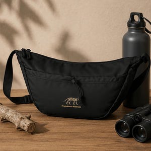 Può includere: Una borsa a tracolla nera a forma di mezzaluna con tracolla regolabile e tasca frontale con cerniera. La borsa presenta una grafica dorata di un tilacino e il testo "THYLACINE museum edition" stampato al centro sulla parte anteriore.