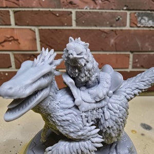 Op de afbeelding: Een gedetailleerd, onbeschilderd miniatuurbeeldje van een kat die op een gevederde dinosaurus rijdt. De kat zit op een zadel op de rug van de dinosaurus. De dinosaurus heeft een lange snuit en gevederde vleugels. Het beeldje is grijs.