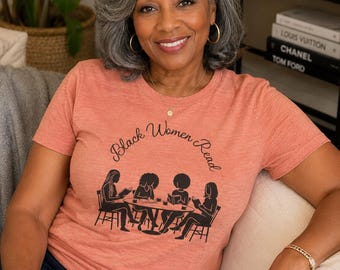 May include: A heathered coral short-sleeve t-shirt featuring a black graphic illustration of four individuals sitting at a round table reading books. The text "Black Women Read" is printed in a black cursive font above the illustration.