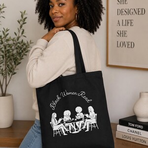May include: A black tote bag featuring a white graphic print of four individuals sitting around a circular table reading books. The text "Black Women Read" is printed in a white cursive font above the illustration of the reading group.