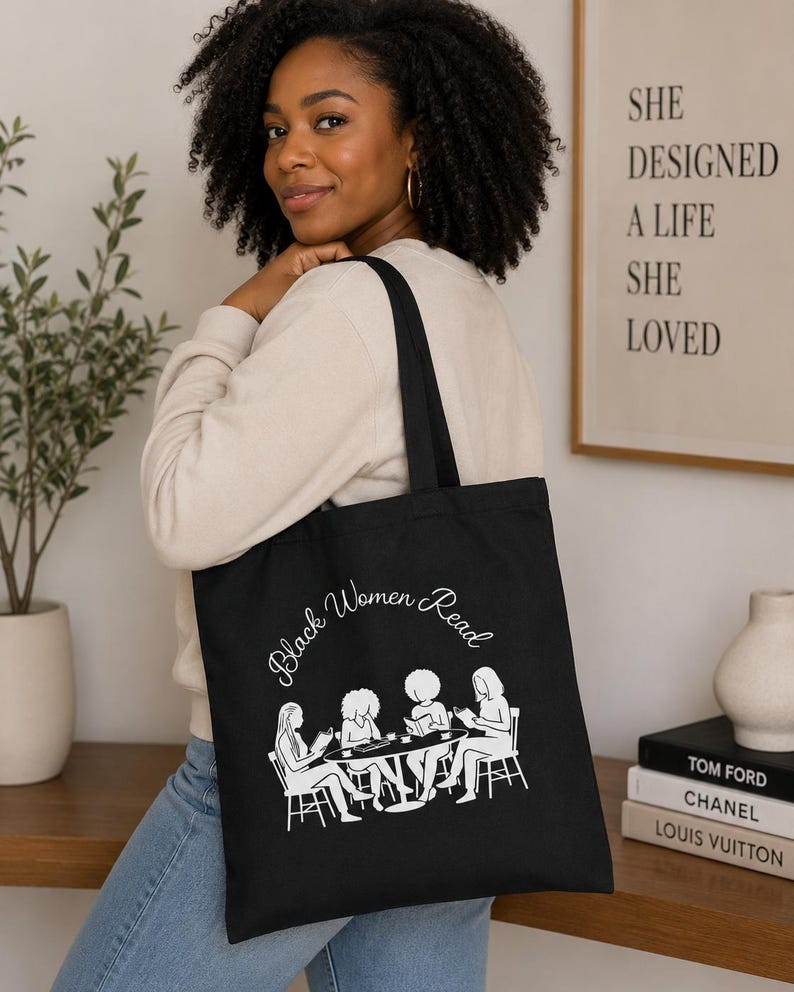 May include: A black tote bag featuring a white graphic print of four individuals sitting around a circular table reading books. The text "Black Women Read" is printed in a white cursive font above the illustration of the reading group.