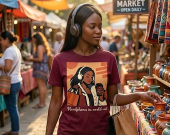 May include: A maroon short-sleeve t-shirt featuring a graphic illustration of a person wearing headphones and holding a shopping bag and a cup, with the text "Headphones in, world out" printed below the graphic in a cursive script font.