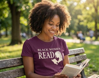 May include: A maroon short-sleeved t-shirt featuring the white text "BLACK WOMEN READ" above a graphic illustration of a person with locs reading an open book. The shirt has a crew neckline and is made of a solid-colored fabric.