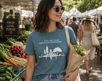 May include: A teal short-sleeved t-shirt featuring a white graphic of a person reading on a park bench with trees and the text Technically Outside MENTALLY ELSEWHERE. The shirt has a crew neckline and a heathered texture throughout the fabric.
