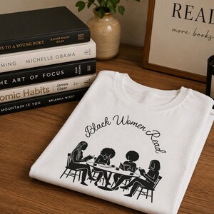 May include: A white t-shirt featuring a black graphic print of four individuals sitting at a table reading books. The text "Black Women Read" is printed in a black cursive font above the illustration of the group gathered around the table.