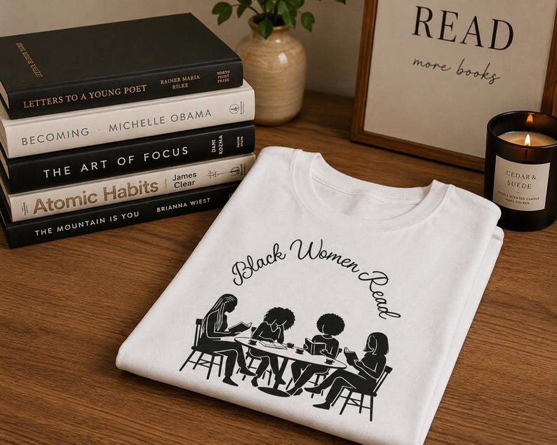 May include: A white t-shirt featuring a black graphic print of four individuals sitting at a table reading books. The text "Black Women Read" is printed in a black cursive font above the illustration of the group gathered around the table.