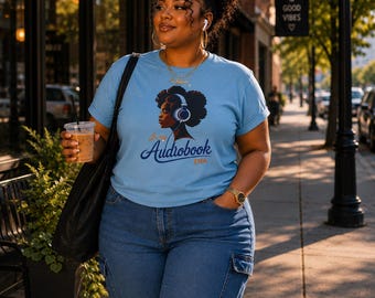 May include: A light blue short-sleeve t-shirt featuring a graphic of a person with an afro wearing headphones. The text on the shirt reads In my Audiobook ERA in dark blue and gold script. The shirt is paired with blue denim cargo pants.