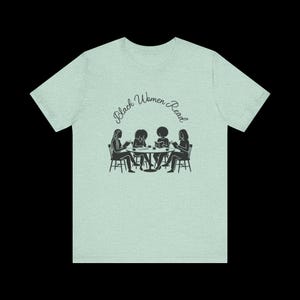 May include: A light green heathered t-shirt featuring a black graphic of four individuals sitting at a round table reading books. Above the illustration, the text Black Women Read is printed in a black cursive font. The shirt has short sleeves.