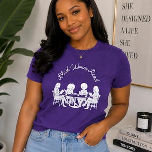 May include: A purple short-sleeved t-shirt featuring a white graphic print of four individuals sitting at a round table reading books. The text "Black Women Read" is printed in a white cursive font above the illustration of the group.