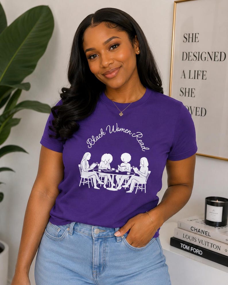 May include: A purple short-sleeved t-shirt featuring a white graphic print of four individuals sitting at a round table reading books. The text "Black Women Read" is printed in a white cursive font above the illustration of the group.