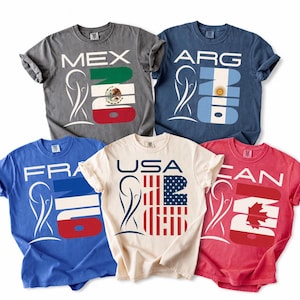May include: Five short-sleeve t-shirts in gray, blue, light blue, cream, and red. Each shirt features a graphic with a stylized trophy icon, the text '26', and country abbreviations: MEX, ARG, FRA, USA, and CAN, incorporating national flag designs.