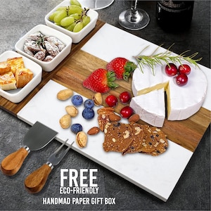 May include: A charcuterie board with white marble and wood sections, featuring cheese, crackers, fruit, and nuts. Includes cheese knives and small white serving dishes. The text "FREE ECO-FRIENDLY HANDMAD PAPER GIFT BOX" is at the bottom.