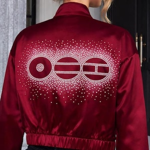 Puede incluir: Una chaqueta bomber de satén rojo oscuro con cuello alto y mangas largas. La parte posterior de la chaqueta está decorada con un patrón grande y complejo de pedrería transparente que forma tres figuras circulares con líneas horizontales.