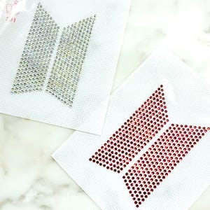 May include: Two iron-on rhinestone transfers featuring the BTS logo shape. One transfer consists of clear, iridescent crystals, while the other is composed of deep red crystals. Both designs are set on white, square adhesive backing sheets.