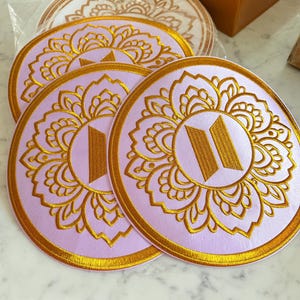 Puede incluir: Tres parches circulares con una base de tela de color morado claro y patrones florales de estilo mandala bordados en oro. Cada parche muestra un logotipo de BTS bordado en oro en el centro. Los bordes tienen un grueso ribete dorado.