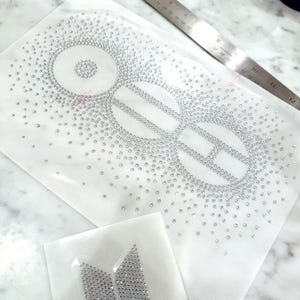 Peut inclure: Une feuille de transfert adhésive transparente avec un motif en strass argentés composé de trois cercles interconnectés et d'un petit écusson trapézoïdal séparé. Une règle métallique montre que le motif mesure environ 30 cm de long.